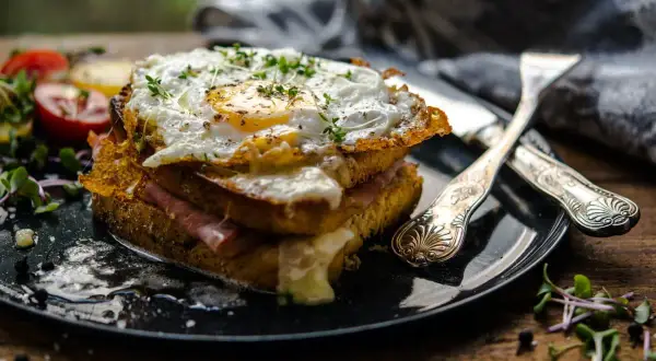 Ein üppiger Croque Monsieur mit Spiegelei, Schinken und geschmolzenem Käse, garniert mit Kräutern und Beilagen.