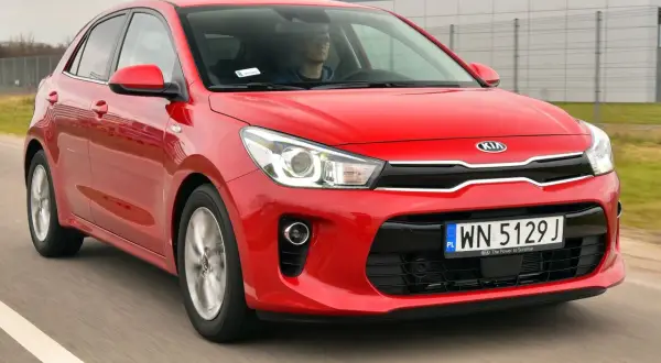 Czerwony kia rio 4 w ruchu, z kierowcą za kierownicą, na tle nowoczesnego budynku.
