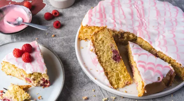 Ein himbeerkuchen, einfach aber extrem lecker, mit rosa Zuckerguss und frischen Himbeeren. Ein Stück ist bereits angeschnitten.