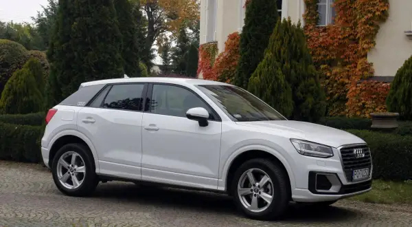 Białe Audi Q2 na brukowanym podjeździe, otoczone zielenią i jesiennymi liśćmi. Idealne do miasta, jak sugerują pierwsze audi q2 opinie.