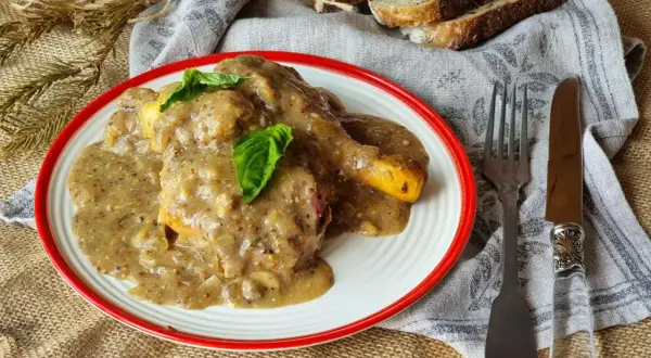 Delicioso pollo a la mostaza cubierto con una cremosa salsa, servido en un plato blanco con borde rojo. Acompañado de pan y cubiertos.