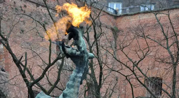Pomnik smoka wawelskiego zieje ogniem na tle ceglanej ściany.