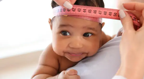 Ein Baby mit dunkler Haut wird der Kopfumfang gemessen. Ein Maßband zeigt etwa 40 cm.
