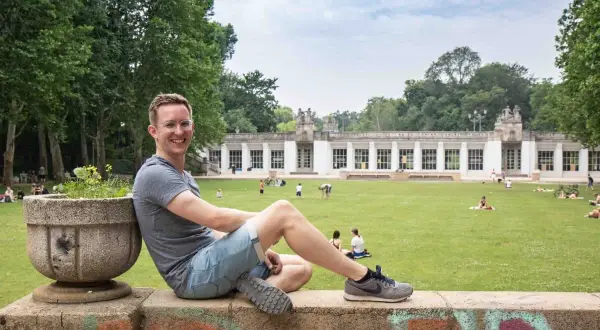 Ein Mann sitzt auf einer Mauer im Park am Stadtbad Charlottenburg Wilmersdorf. Menschen entspannen sich auf der Wiese vor dem Gebäude.