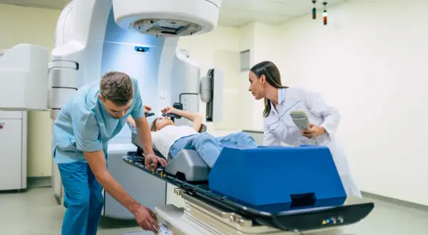 Ein Patient liegt auf einer Liege, während ein Techniker und eine Ärztin eine perkutane Strahlentherapie vorbereiten.