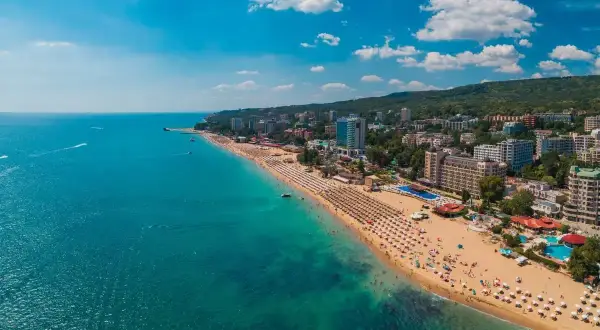 Słoneczna plaża w Złote Piaski, z mnóstwem leżaków i turystów. W tle widać hotele i zielone wzgórza. Idealne miejsce na wakacje, niedaleko lotnisko.