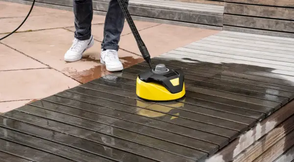 Osoba myje drewniany taras na balkonie w bloku za pomocą żółtej myjki ciśnieniowej.