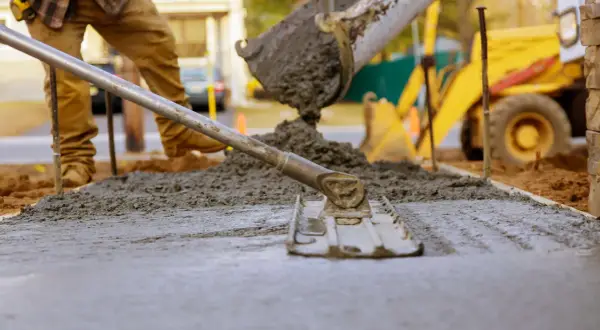 Pracownik wylewa beton na chudziaka, używając kielni do wyrównania powierzchni. W tle widać koparkę.