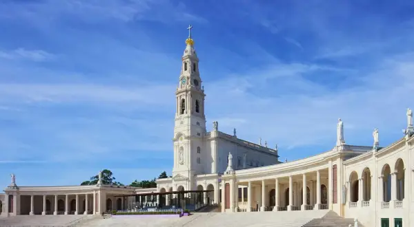 Biała bazylika z wieżą i złotą koroną na szczycie, otoczona kolumnadą. To Sanktuarium Matki Bożej Różańcowej w Fatimie, miejsce pielgrzymek.