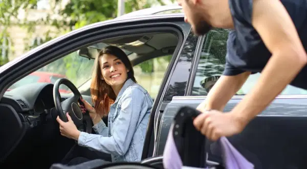Una donna disabile sorride al volante mentre un uomo aiuta a caricare la sua sedia a rotelle in auto.