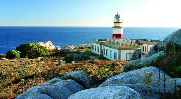Faro en las islas Galicia, con el mar azul de fondo y rocas cubiertas de vegetación.
