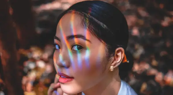 Un occhio a mandorla cattura la luce arcobaleno, riflessi colorati danzano sul suo viso mentre guarda pensierosa.