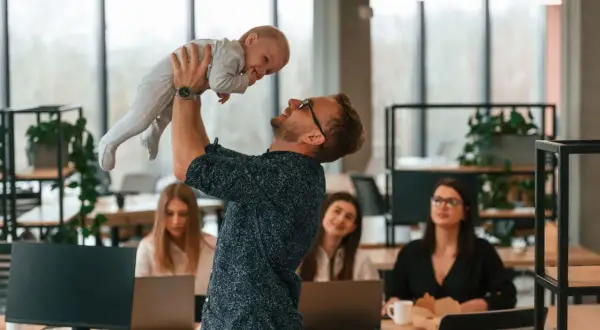 Szczęśliwy ojciec podrzuca dziecko w biurze, podczas gdy koleżanki pracują. Czy urlop wychowawczy wlicza się do stażu pracy?