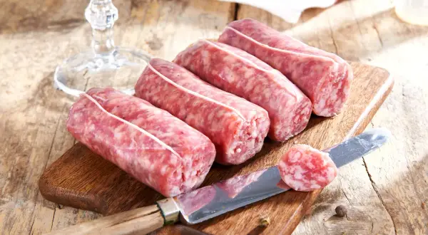 Frische Salsiccia-Würste liegen auf einem Holzbrett. Ein Messer mit einem Stück Wurst zeigt, was ist Salsiccia: grob gehacktes Schweinefleisch, gewürzt und in Darm gefüllt.