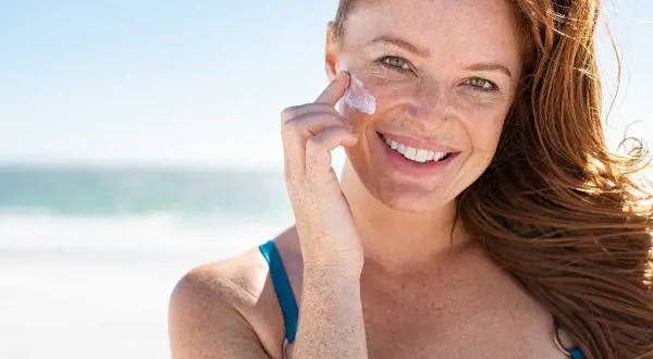 Donna sorridente con lentiggini applica crema solare sul viso in spiaggia. Si può mettere il fondotinta sopra la crema solare per un look perfetto.