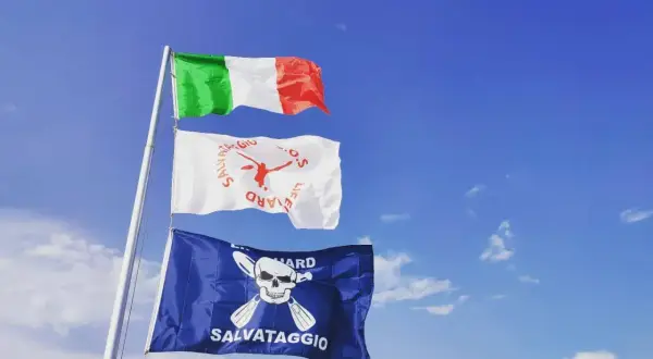 Bandiera azzurra con croce bianca, simbolo di salvataggio, sventola accanto alla bandiera italiana sotto un cielo azzurro.