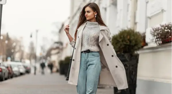 Styling Mom Jeans: Ein lässiger Look mit Strickpullover, Trenchcoat und Perlenschmuck auf der Straße.