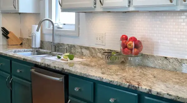 A kitchen with a white subway tile backsplash, granite countertops, and teal cabinets. A bowl of apples sits on the counter.