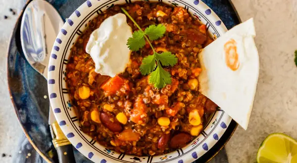 Herzhaftes Chili sin Carne mit Quinoa, Bohnen und Mais, garniert mit Sauerrahm und Koriander. Dazu eine Tortilla-Ecke und Limettenspalten.