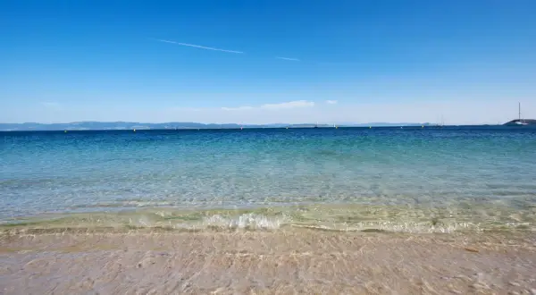 Aguas cristalinas en la playa milide, con veleros navegando en el horizonte bajo un cielo azul intenso.