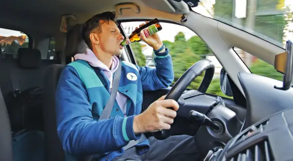 Mężczyzna za kierownicą pije piwo. Czy pasażer może pić alkohol? To niebezpieczne zachowanie.
