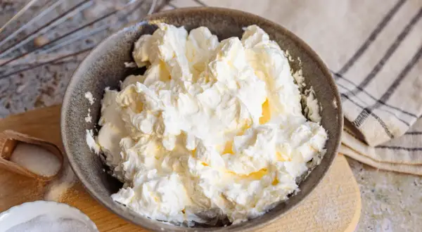 Schüssel mit luftiger, weißer italienischer Buttercreme, bereit für süße Kreationen. Daneben Zucker und ein Schneebesen.