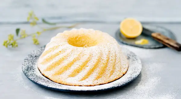 En vackert pudrad sockerkaka med citron, serverad på en mörk tallrik med en citronskiva och en kniv i bakgrunden.