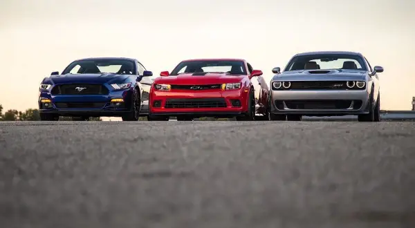 Trzy potężne muscle cary: niebieski Mustang, czerwony Camaro i srebrny Challenger, stoją obok siebie na tle zachodzącego słońca.