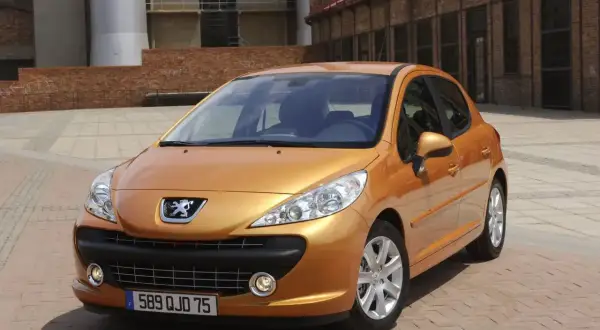 Pomarańczowy Peugeot 207 1.4 benzyna zaparkowany na placu z ceglaną posadzką, w tle budynki przemysłowe.