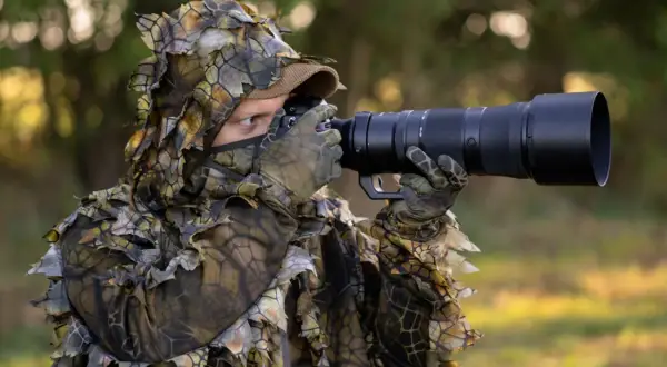 Osoba w kamuflażu z liśćmi czeka na ptaki, gotowa do fotografowania.