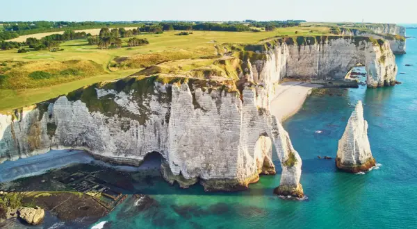 Wapienne klify nad kanałem La Manche, z łukiem skalnym i iglicą. Na szczycie klifów zielone pola golfowe.