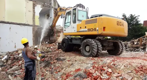 Robot budowlany i pracownik z wężem wodnym podczas rozbiórki. Szacowany koszt wyburzenia domu 100m2.