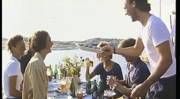 En glad grupp njuter av en sommarmåltid vid havet, en scen som påminner om en klassisk pripps blå reklam.