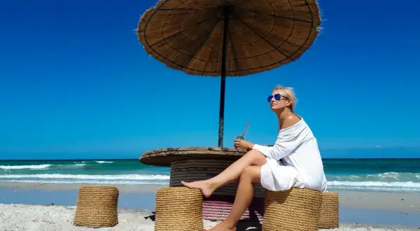 Kobieta w białej sukience cieszy się drinkiem pod parasolem na plaży. Idealne tunezja wakacje.