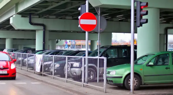 Czerwony samochód zbliża się do parkingu, gdzie stoi znak b2. Zielony samochód zaparkowany jest pod wiaduktem.