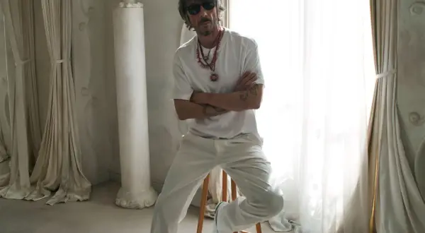Pierpaolo Piccioli, con gafas de sol y collares llamativos, posa con los brazos cruzados en un entorno elegante con cortinas y columnas.