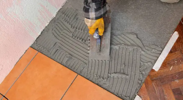 Ręka w żółtej rękawicy rozprowadza klej na podłodze za pomocą kielni. To kluczowy etap, jak nakładać klej na płytki, by zapewnić trwałe połączenie.