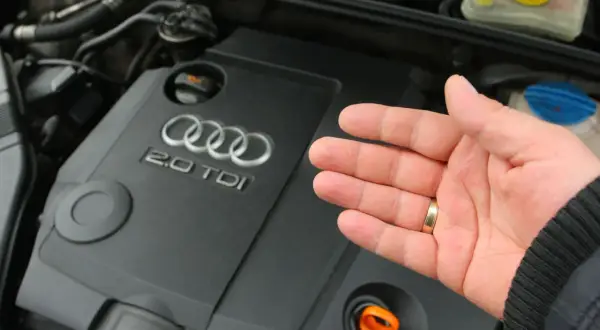 Dłoń pokazuje silnik Audi 2.0 TDI. Widoczny jest pomarańczowy bagnet do sprawdzania poziomu oleju, a obok niego korek, pod którym może znajdować się masło pod korkiem oleju.