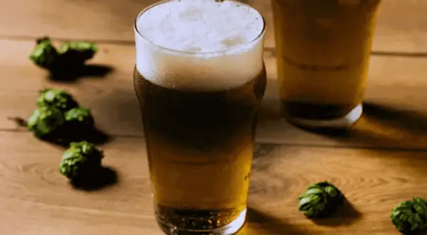 Bicchiere di birra dorata con schiuma bianca, circondato da luppoli verdi su un tavolo di legno. Perfetta per una dual IPA.