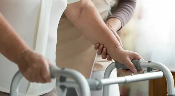 Una persona anziana, con l'aiuto di un'assistente, si appoggia a un deambulatore, mostrando la difficoltà nel riuscire a camminare all'improvviso.