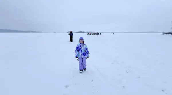 Dziewczynka w kolorowym kombinezonie na tle zamarzniętego jeziora. W oddali widać grupę ludzi. To niezapomniana laponia wycieczka.
