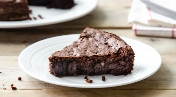 Una fetta di torta al cioccolato senza farina e burro, densa e umida, su un piatto bianco.