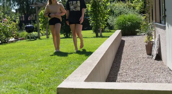Två flickor går på en gräsmatta bredvid en nybyggd mur i en solig trädgård.