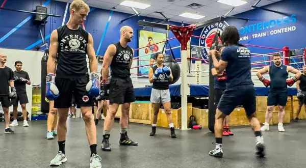 Boxers in a gym, wearing gloves and training gear, practice their moves. One person is hitting a punching bag, while others watch and wait for their l step.