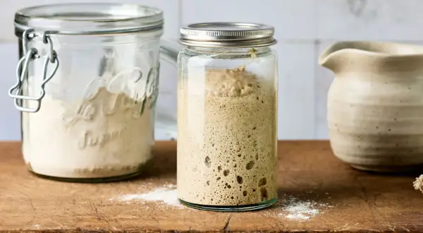 Ein Glas mit blubberndem Sauerteig, bereit zum Backen. Daneben ein Glas mit Mehl, um den Sauerteig anzusetzen.