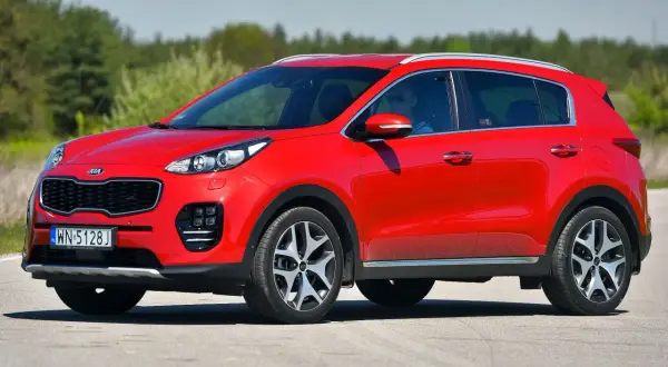 Czerwony Kia Sportage IV z kierowcą w okularach przeciwsłonecznych jedzie po drodze, otoczony zielenią.
