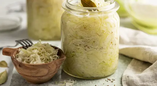 Frisches Sauerkraut im Glas, bereit für ein leckeres Sauerkraut Rezept. Ein kleiner Holzlöffel mit Sauerkraut und Lorbeerblätter liegen daneben.