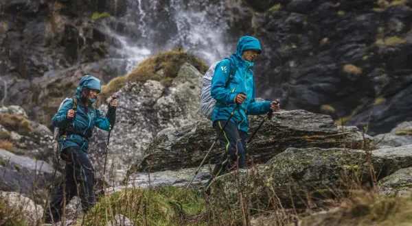 Dwie osoby w turkusowych kurtkach w góry, z kijkami trekkingowymi, idą obok wodospadu.
