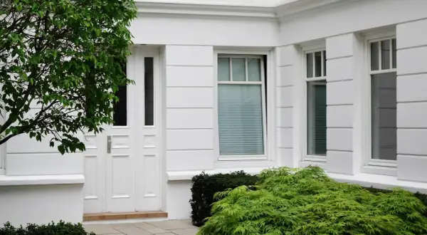 Białe drzwi i okna eleganckiego domu, otoczone bujną zielenią. Drzewo z liśćmi rzuca cień na fasadę, a krzewy dodają uroku.