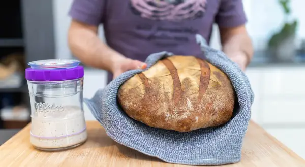 Un panadero muestra un pan artesanal recién horneado junto a un frasco de masa madre activa, listo para empezar a hacer masa madre.